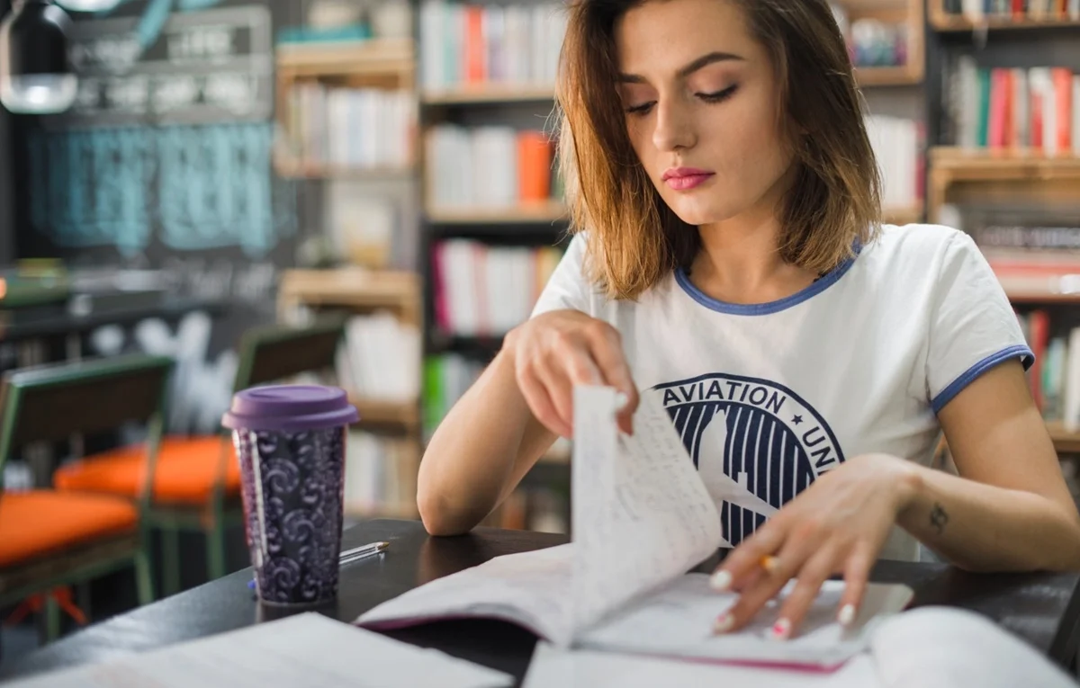 imagem de uma jovem sentada na mesa de uma biblioteca folheando o caderno.
